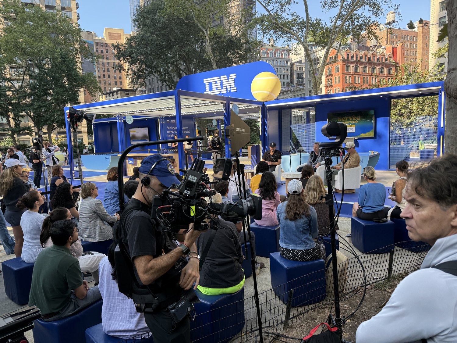 The live Served taping in Madison Square Park: a blue IBM pop-up with a giant tennis ball, the panel on stage, an audience seated on blue blocks, and a camera operator shooting from the foreground.