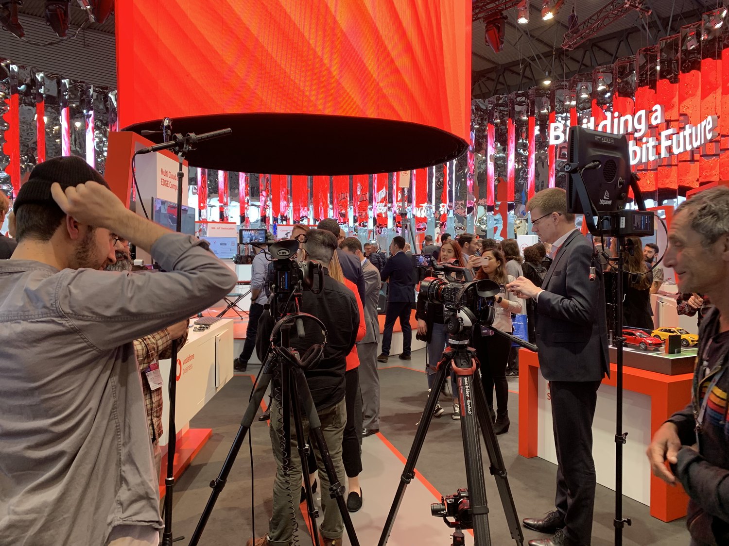 A live event floor under red LED walls: several camera crews shooting interviews at once in a crowded show stand.